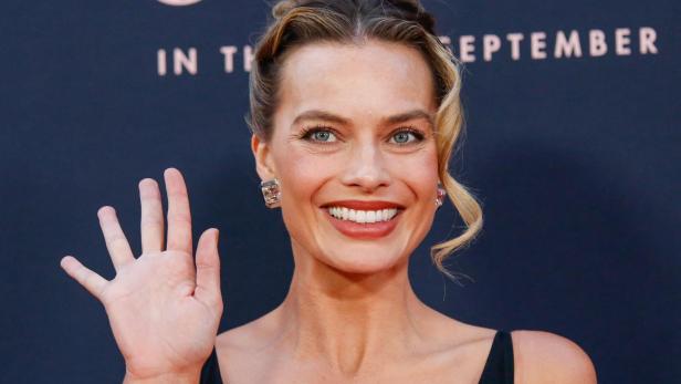 Eine Frau mit hochgesteckten Haaren lächelt und winkt mit der rechten Hand vor dunklem Hintergrund.