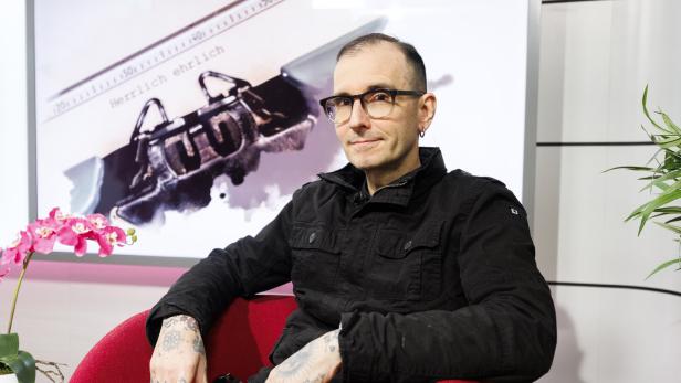 Ein Mann mit Brille und Tattoos sitzt in einem roten Sessel, im Hintergrund eine Schreibmaschine und eine pinke Orchidee.