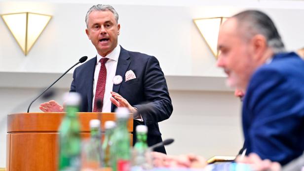 Ein Mann im Anzug spricht an einem Rednerpult, im Vordergrund sitzen weitere Personen mit Flaschen auf dem Tisch.