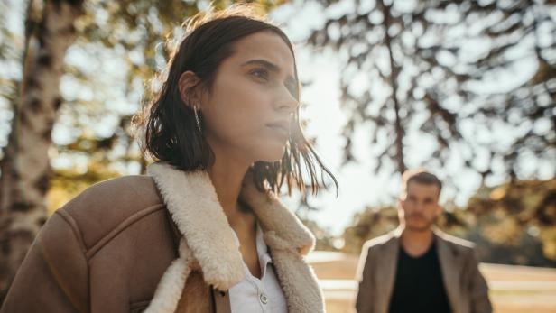 Eine Frau mit brauner Jacke und Fellkragen schaut besorgt aus, ein Mann steht im Hintergrund.