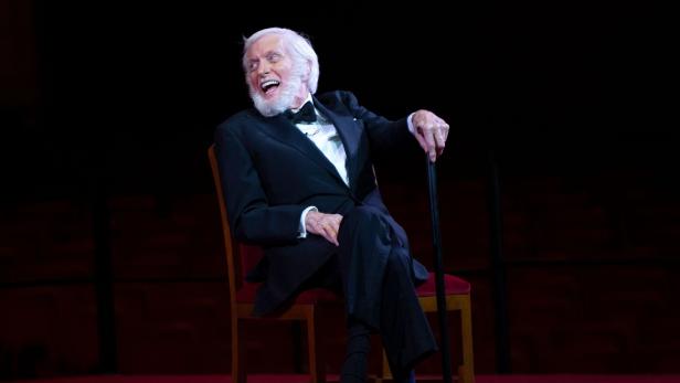 Ein älterer Mann im Smoking sitzt lachend auf einem Stuhl und hält einen Gehstock auf einer Bühne.