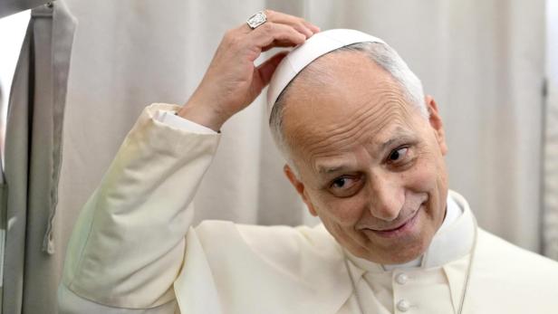 Der Papst in seinem weißen Ornat fasst sich an seine Mütze, während er den Kopf etwas schrägt hält.