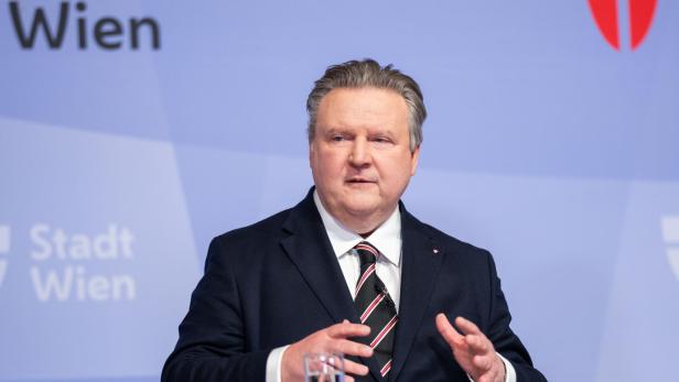 Ein Mann im Anzug spricht gestikulierend vor einem hellblauen Hintergrund mit dem Schriftzug „Stadt Wien“ und einem roten Wappen.