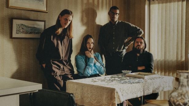 Die Band Dry Cleaning, ernst an einem Tisch in einer altmodisch möblierten Wohnung sitzend und stehend