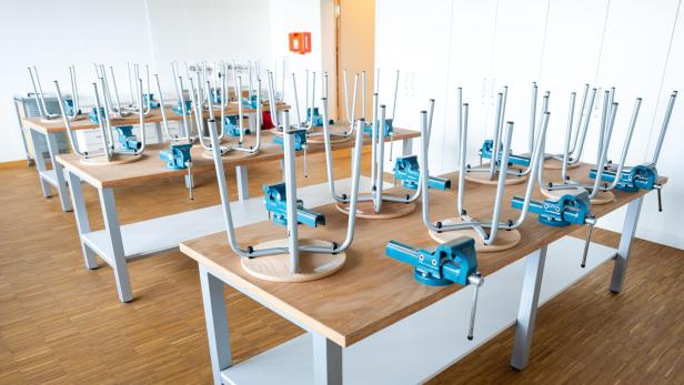 Mehrere Werkbänke mit Schraubstöcken, auf denen umgedrehte Hocker stehen, in einem hellen Raum mit Holzfußboden.