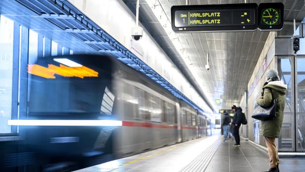 U-Bahn-Station mit vorbeirasendem Zug, wartenden Fahrgästen und Anzeige „Karlsplatz“.