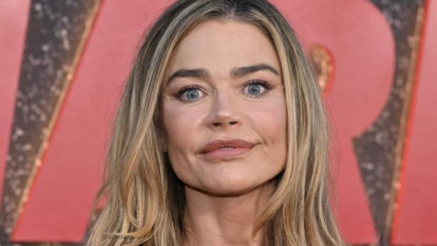 Denise Richards trägt ein schwarzes, glitzerndes Oberteil und eine Halskette mit Kreuzanhänger vor einem roten Hintergrund.