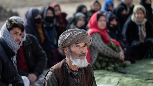 Ein älterer Mann mit Turban sitzt im Vordergrund, im Hintergrund sitzen mehrere Frauen und Männer mit Kopftüchern.