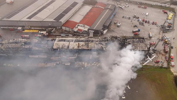Rauch steigt aus der zerstörten Keksfabrik von Violanta auf, während zahlreiche Einsatzfahrzeuge und Feuerwehrleute am Unglücksort arbeiten.