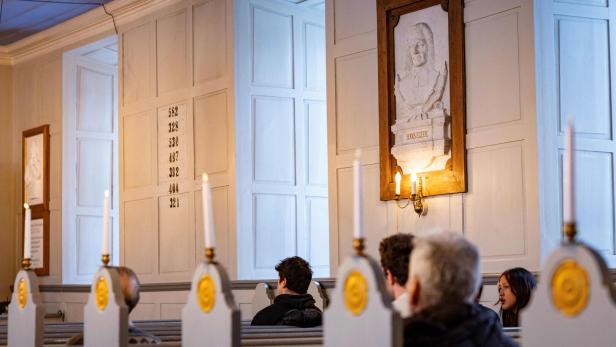 In einer Kirche sitzen Menschen in Bänken mit Kerzen, an der Wand hängt ein Relief von Hans Egede.