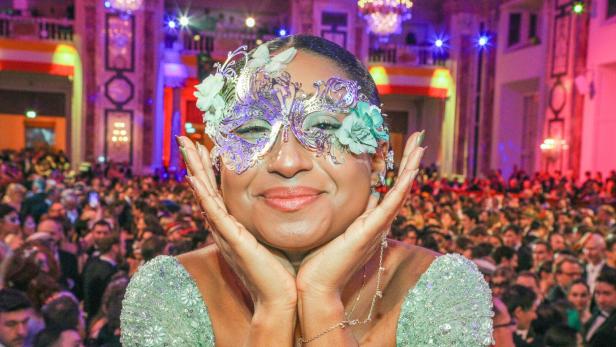 Eine Frau mit verzierten Maske und grünem Kleid lächelt, während sie von einer festlich gekleideten Menschenmenge umgeben ist.