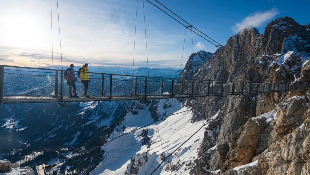 Zwei Personen stehen auf einer Hängebrücke hoch über einer verschneiten Berglandschaft bei Sonnenschein.