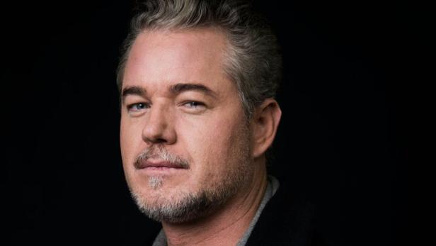 Eric Dane mit grau meliertem Haar und Bart trägt einen dunklen Mantel über einem grauen Hoodie und einem weißen T-Shirt.