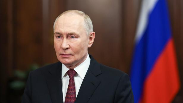 Wladimir Putin trägt einen dunklen Anzug mit Krawatte vor der russischen Flagge.