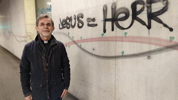 Ein Mann mit Priesterkragen steht vor einer Wand mit dem Graffiti „JESUS = HERR“ und kleinen grünen Herzen.