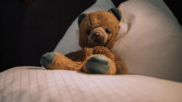 Abgenutzter Teddybär sitzt allein auf einem weißen Kopfkissen im Bett.