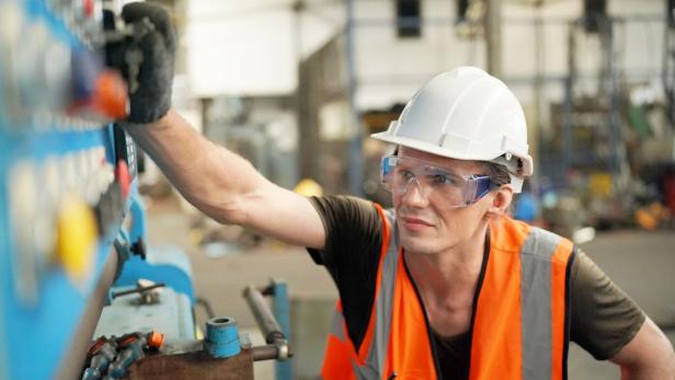 Eine Person mit Schutzhelm und Warnweste bedient konzentriert eine Maschine in einer Werkhalle.