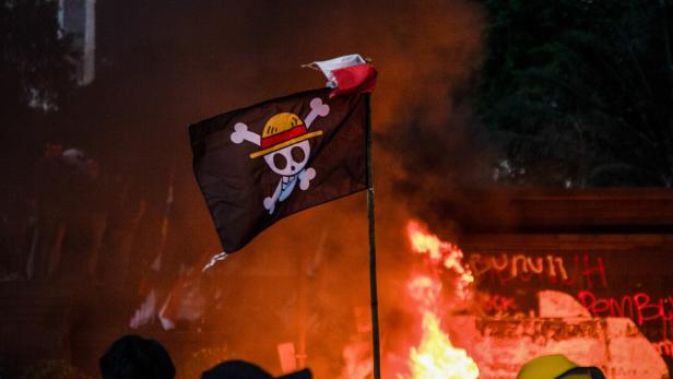 Menschenmenge vor einem großen Feuer, darüber eine schwarze Piratenflagge mit Strohhut und gekreuzten Knochen.
