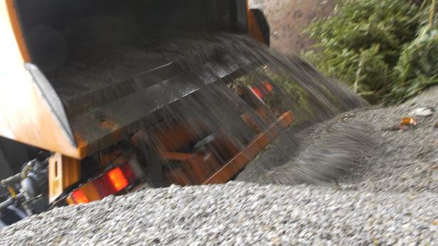 Ein Lastwagen kippt eine große Menge Kies auf einen Haufen neben einigen grünen Ästen ab.