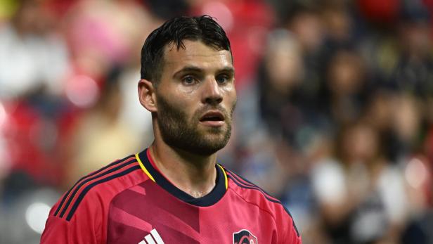 Eduard Löwen mit Bart trägt ein rotes Trikot mit Sponsorenlogos vor verschwommenem Publikum.