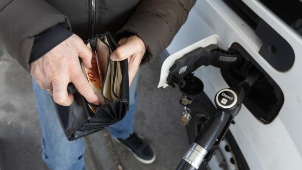 Eine Person hält an einer Tankstelle ein Portemonnaie mit Euro-Scheinen in der Hand, während sie ihr Auto mit Diesel betankt.