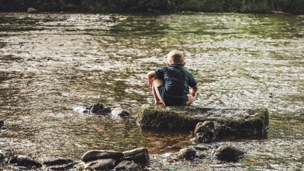 Ein Junge spielt an einem Fluss und wird von hinten fotografiert.