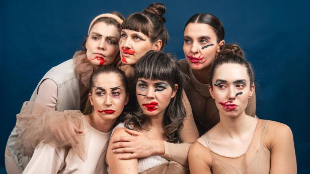 Sechs Frauen mit verschmiertem, künstlerischem Make-up posieren eng beieinander vor blauem Hintergrund.