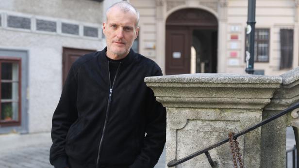 Ein Mann mit grauem Haar und Bart steht in schwarzer Jacke an einer steinernen Brüstung in einer Altstadtgasse.