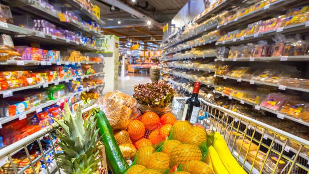 Ein Einkaufswagen voll mit frischem Obst, Gemüse, Brot und einer Weinflasche steht in einem Supermarkt.
