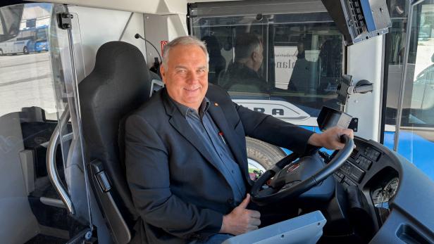 Ein lächelnder Mann im Anzug sitzt am Steuer eines Busses.