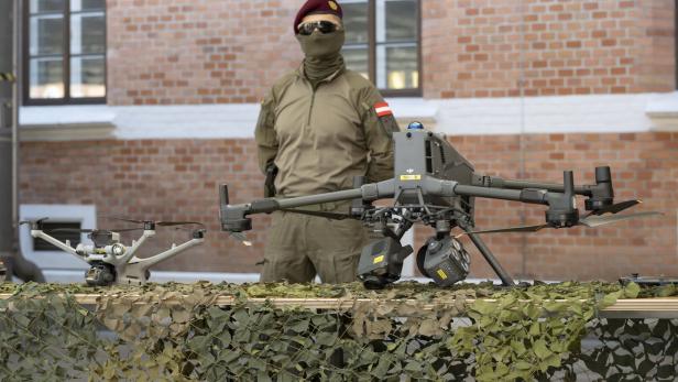 Ein Soldat in Uniform steht hinter zwei Überwachungsdrohnen auf einem mit Tarnnetz bedeckten Tisch.