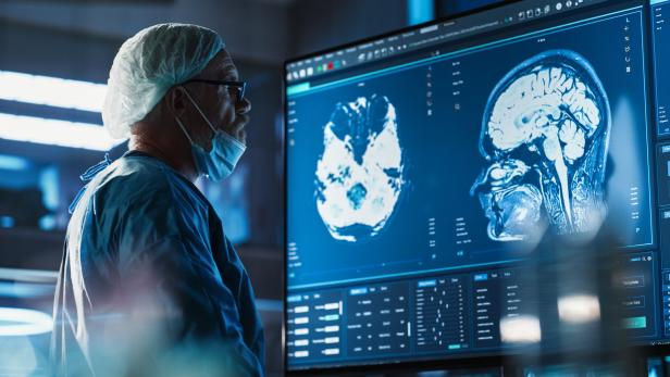 Medical Hospital Research Laboratory: Caucasian Male Neurosurgeon Looking At TV Screen With Brain MRI Scans Of Patient, Analyzing, Preparing For Complicated Neurosurgery, Wearing Surgical Gown.