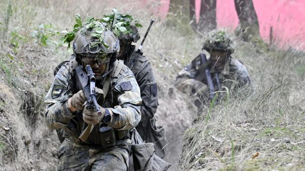 Drei Soldaten in Tarnuniform mit Gewehren bewegen sich durch einen Graben im Wald, im Hintergrund ist rosa Rauch zu sehen.