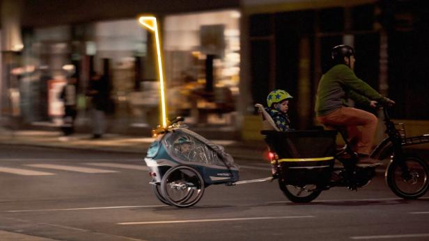Ein Radfahrer fährt bei Dunkelheit mit einem Kind im Lastenrad und einem beleuchteten Fahrradanhänger über eine Straße.
