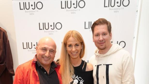 Drei Personen stehen lächelnd nebeneinander vor einer Werbewand mit dem Schriftzug „LIU·JO“.