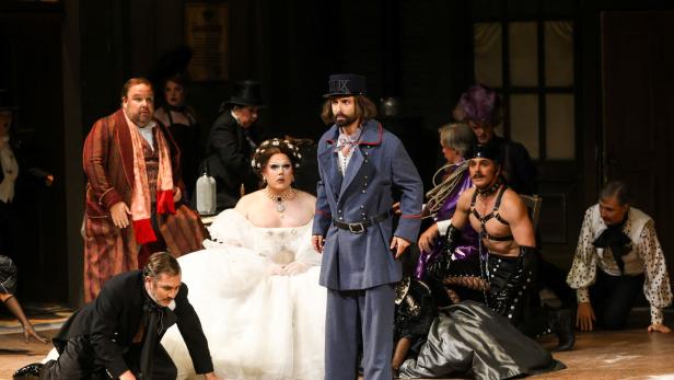 Mehrere Menschen in aufwendigen, historischen und extravaganten Kostümen stehen und knien in einer Theaterszene.