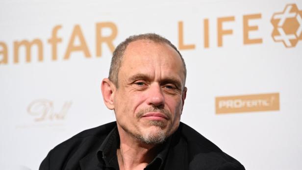 Mann mit kurzer Glatze und Spitzbart vor weißem Event-Hintergrund mit amfAR LIFe-Logo.