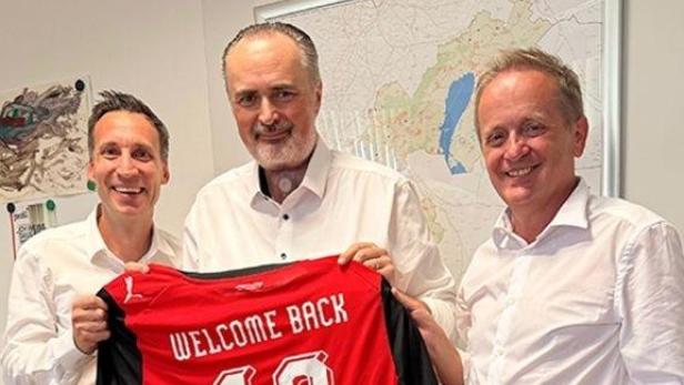 Drei Männer in einem Büro halten ein rotes Trikot mit der Aufschrift „Welcome Back 10 Dosko“.