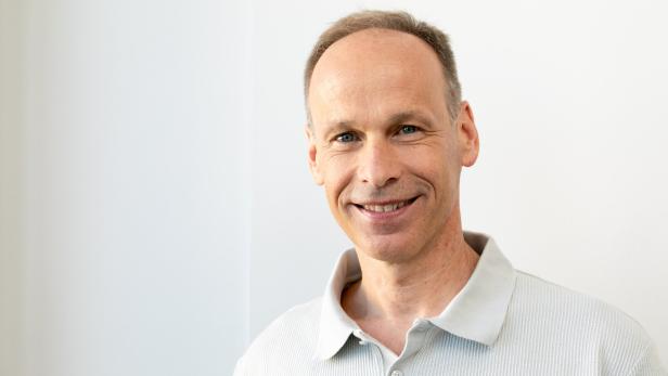 Ein lächelnder Mann mit kurzem Haar trägt ein helles Poloshirt vor einem weißen Hintergrund.