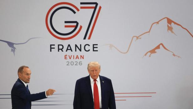 Donald Trump steht vor einem G7-Gipfel-Hintergrund in Évian, während ein Mann neben ihm in eine Richtung deutet.