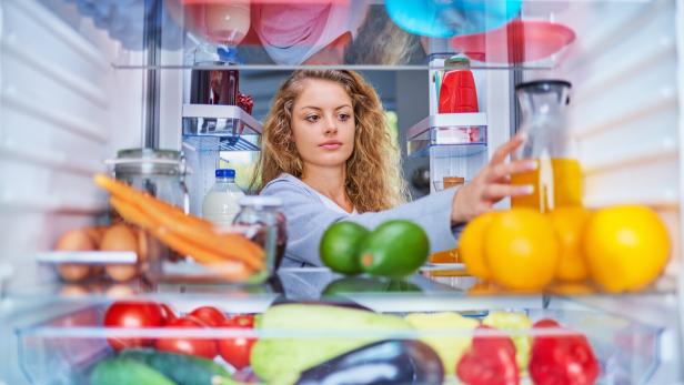 Eine junge Frau mit lockigem Haar greift aus der Perspektive des Kühlschrankinneren nach einem Glas Saft zwischen frischem Obst und Gemüse.