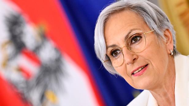 Ältere Frau mit Kurzhaarschnitt und Brille vor unscharfem rot-blauem Hintergrund.