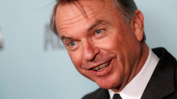 Der Schauspieler Sam Neill lächelt vor einem hellblauen Hintergrund.