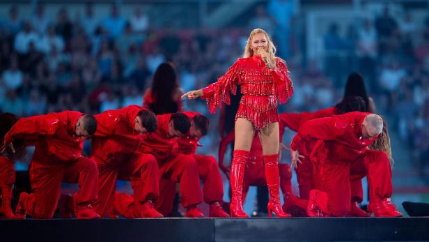 Helene Fischer singt in einem roten Fransen-Outfit auf einer Bühne, umgeben von knienden Tänzern in roter Kleidung.