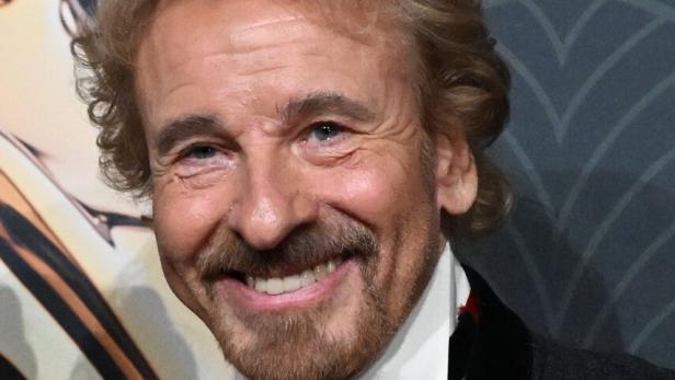 Thomas Gottschalk lächelt in einem dunklen Anzug mit weißem Hemd und schwarzer Fliege vor einem gemusterten Hintergrund.