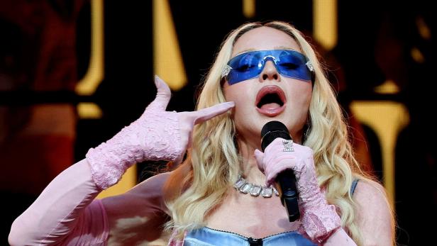 Madonna singt in ein Mikrofon, trägt eine blaue Sonnenbrille, ein blaues Korsett sowie rosa Spitzenhandschuhe und gestikuliert mit der Hand am Gesicht.