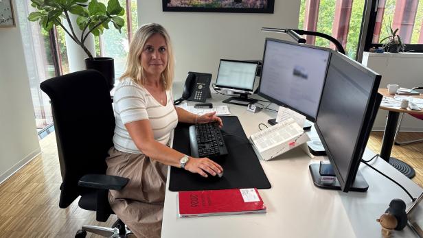 Frau sitzt an einem modernen Schreibtisch im Büro mit zwei Monitoren und Tastatur, umgeben von Pflanzen und Tageslicht.