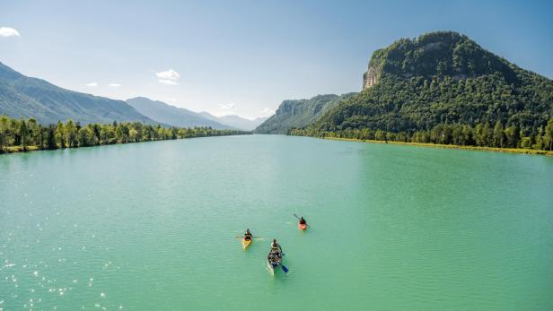 Drei Boote mit Menschen paddeln auf einem türkisfarbenen Fluss vor einer bewaldeten Bergkulisse.