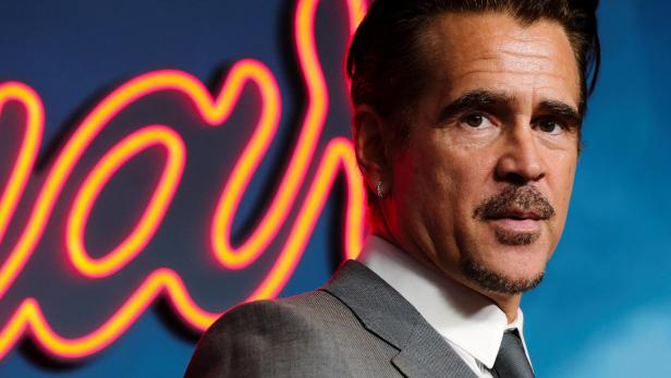 Colin Farrell trägt einen grauen Anzug vor einem blauen Hintergrund mit leuchtend orangefarbenen Neonbuchstaben.