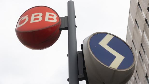 Ein rotes ÖBB-Logo und ein blaues Schild mit weißem Zickzack-Symbol an einem Mast vor einem Gebäude.